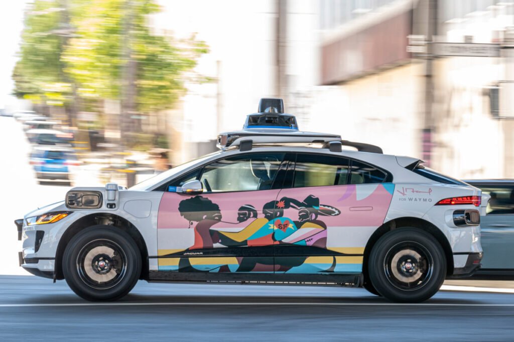 Carro autônomo Waymo branco, modelo Jaguar I-Pace, com arte lateral colorida, em movimento numa rua urbana.