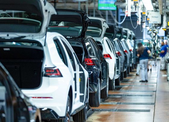 Linha de montagem de carros em fábrica, com veículos brancos e pretos, porta-malas abertos e trabalhadores.