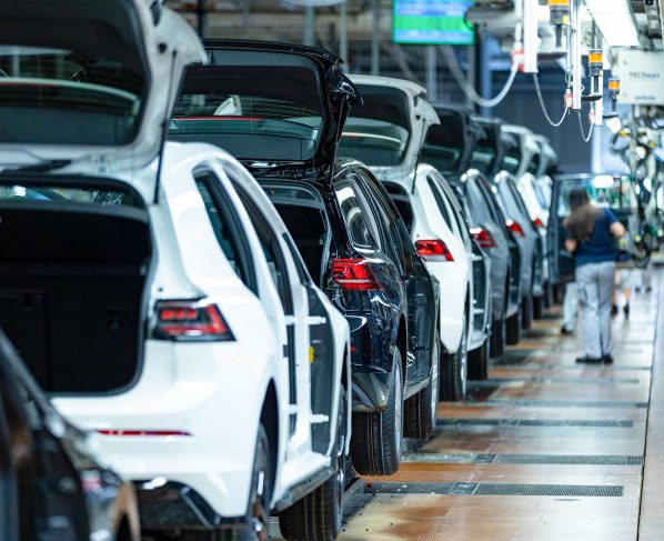 Linha de montagem de carros em fábrica, com veículos brancos e pretos, porta-malas abertos e trabalhadores.