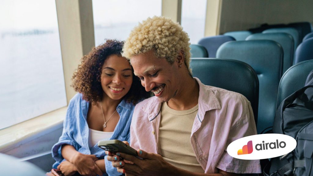 Casal sentado em transporte olha um smartphone juntos enquanto sorri. A imagem inclui o logotipo da Airalo no canto inferior direito.